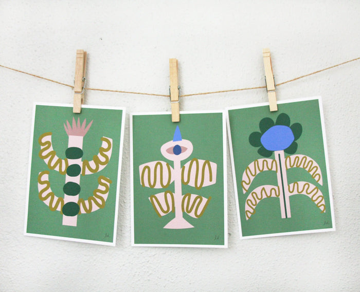 Three abstract bird and flower prints in green tones hanging on a string with clothespins against a white wall